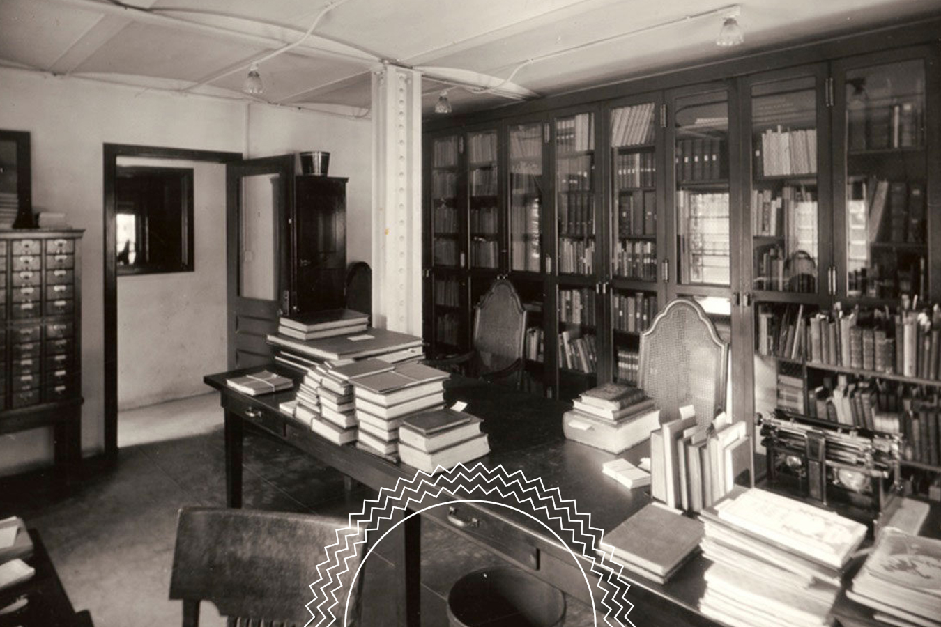 A vintage library room with bookshelves, glass cabinets, and desks stacked with books. There are several chairs and a filing cabinet against the wall.