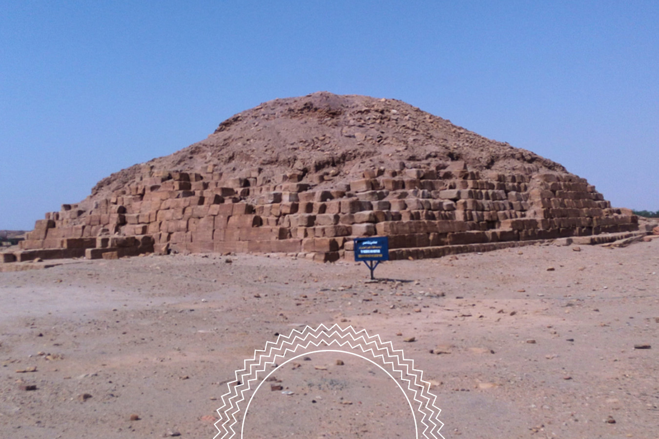 A stepped stone pyramid with a weathered top stands in a desert landscape under a clear blue sky. A blue informational sign is placed in front of the structure.