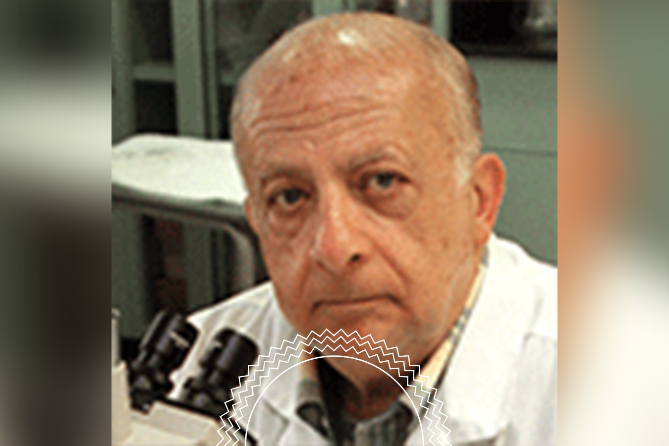 An older man in a lab coat sits in a laboratory next to a microscope, looking toward the camera. Shelves and lab equipment are visible in the background.