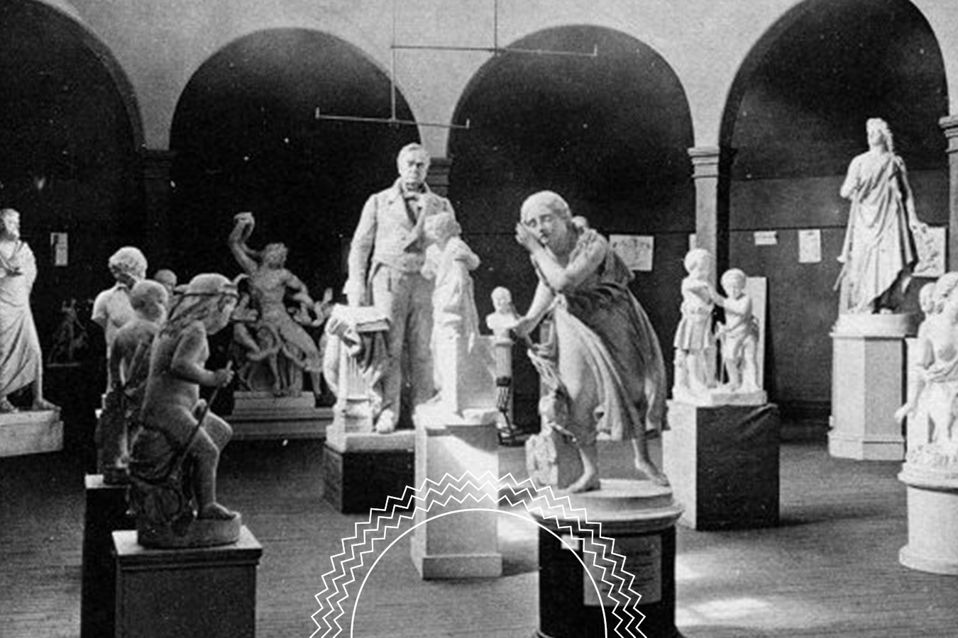 Black and white photo of a museum room displaying multiple classical statues, including standing and seated figures, under arches in the background.