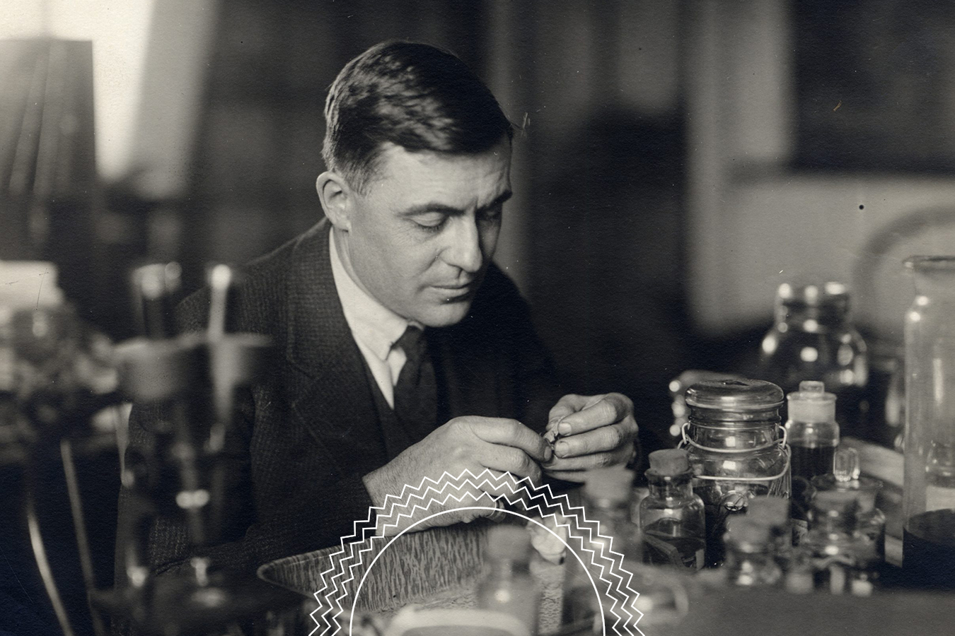 A man in a suit sits at a cluttered laboratory desk, handling a small object, surrounded by glass jars and scientific equipment.