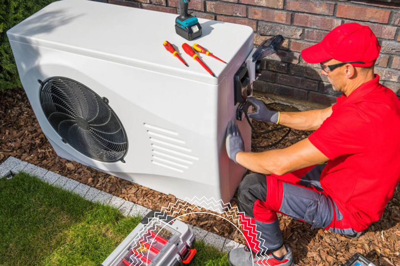 A technician in red uniform installs or repairs a large outdoor air conditioning unit next to a brick wall, with tools and a toolbox nearby.