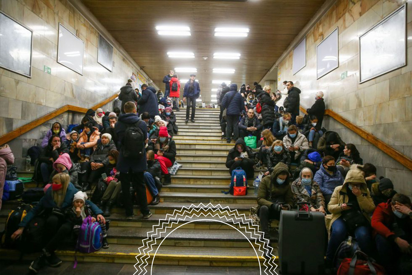 A large group of people, some wearing masks, sit and stand on steps in an underground space, with bags and luggage around them.