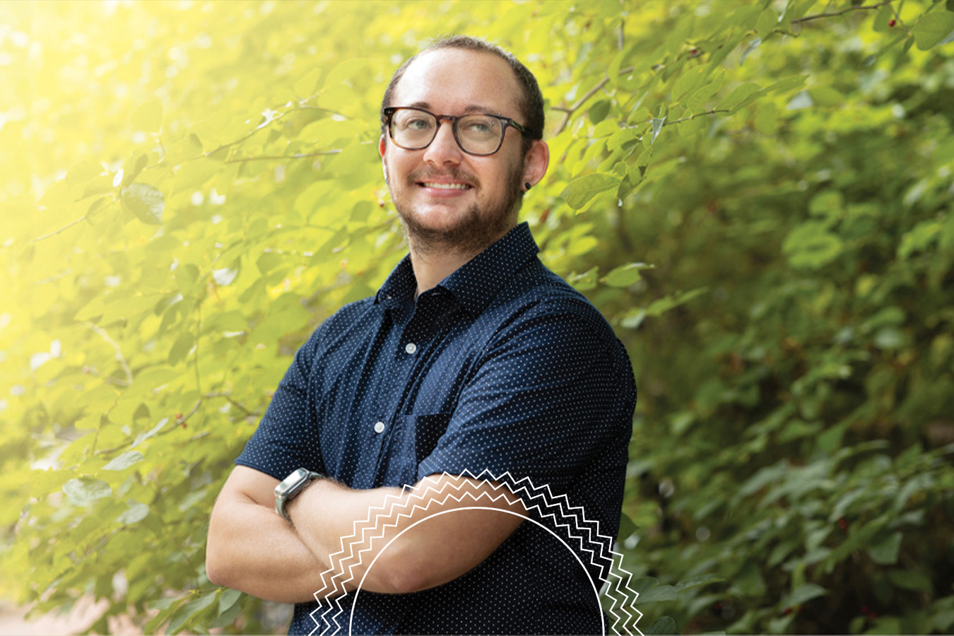 A person wearing glasses and a short-sleeve button-up shirt stands outdoors, smiling with arms crossed in front of leafy green foliage.