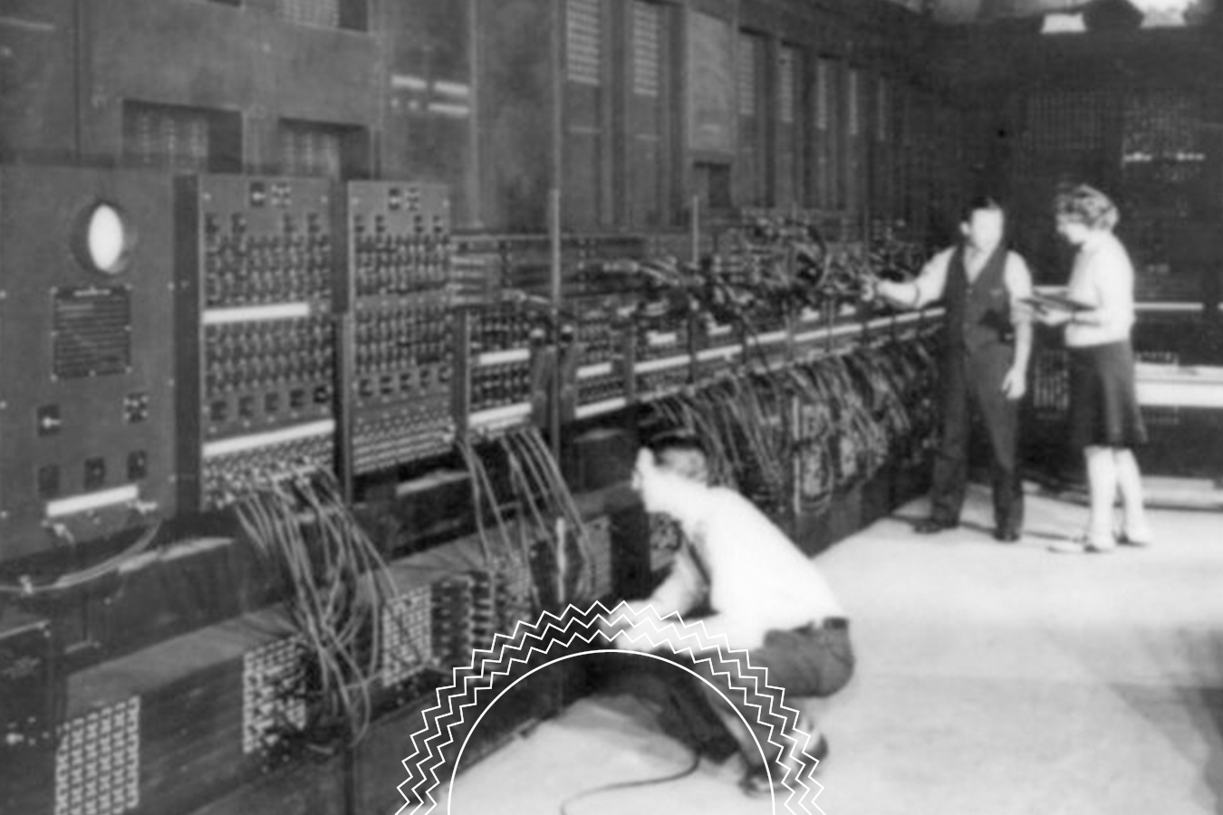 Three people operate a large, early computer with numerous cables and switches in a room filled with equipment.