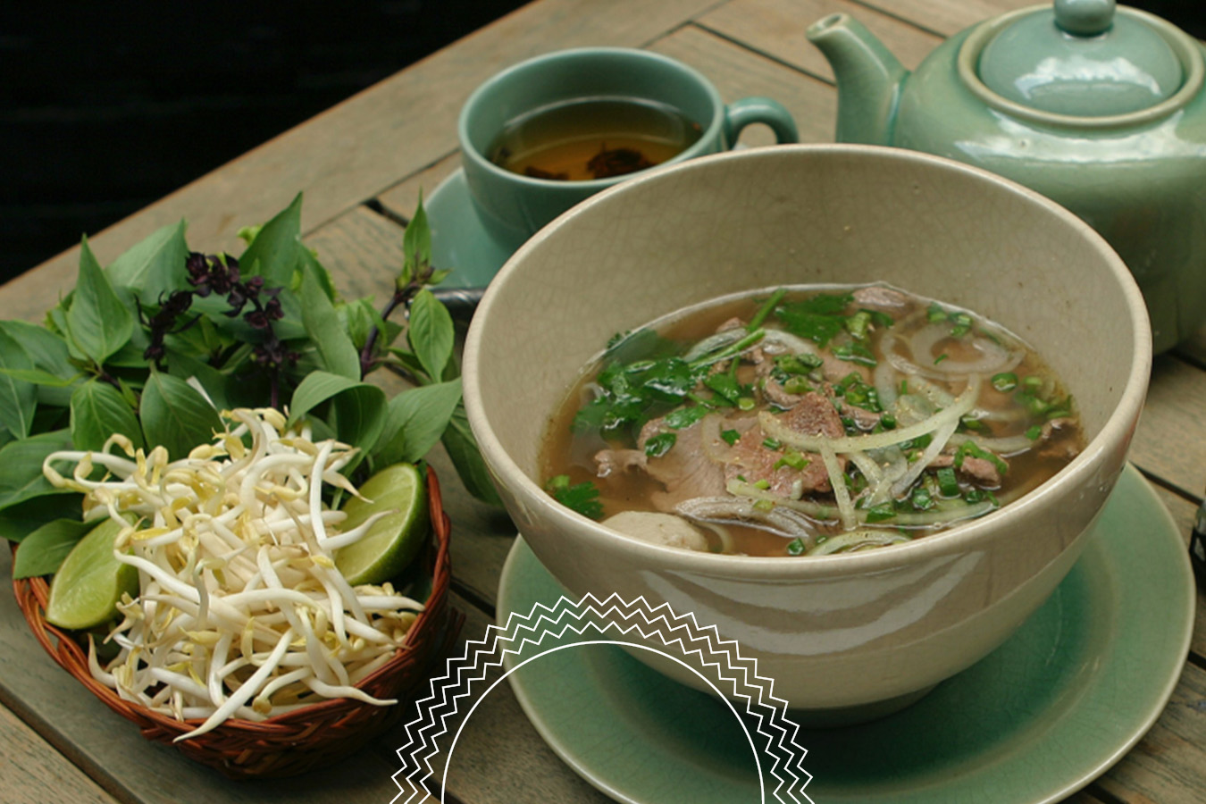 A bowl of pho with noodles, beef, and herbs is served with a side of bean sprouts, lime, basil, and a cup of tea on a wooden table.