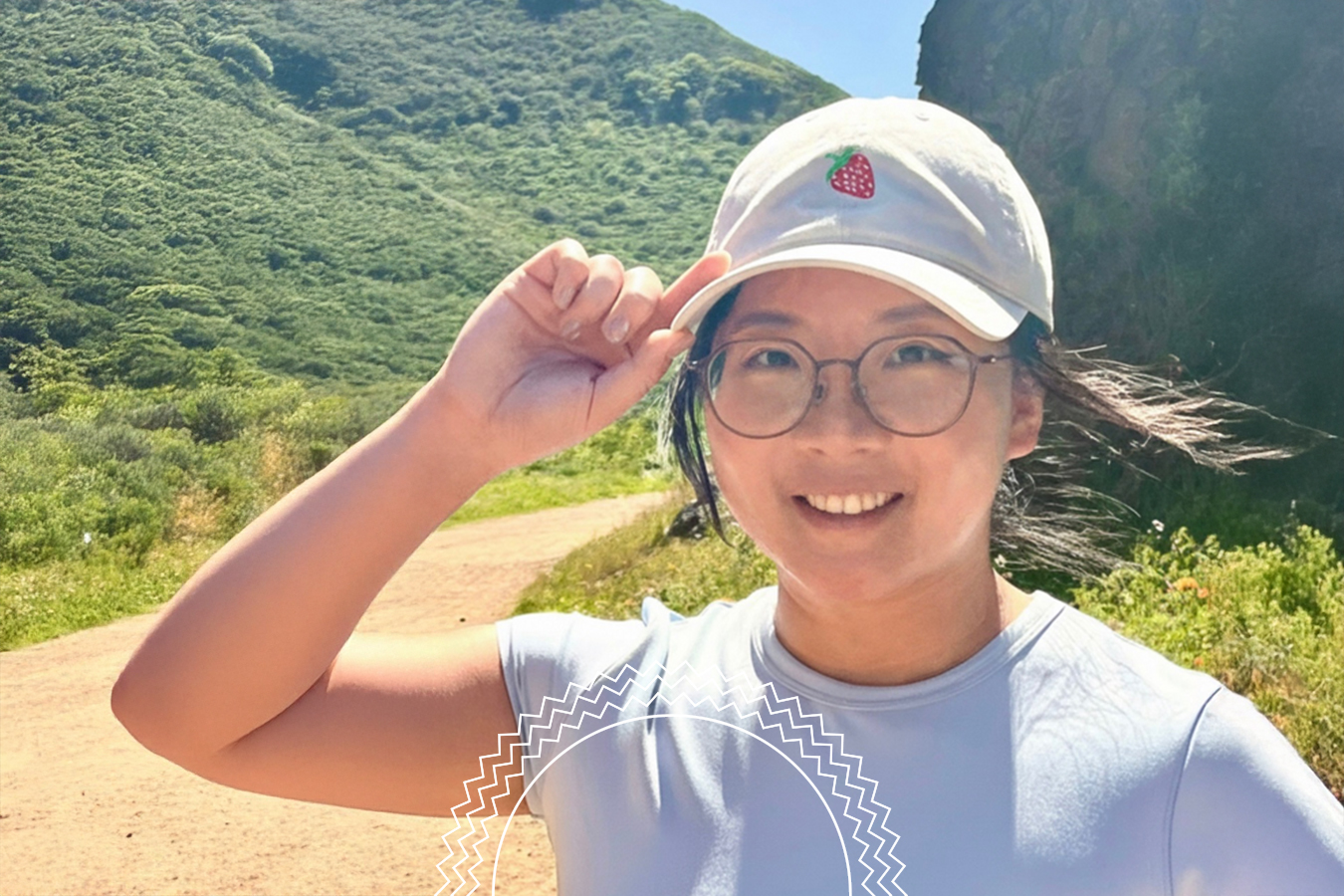 Person wearing glasses and a strawberry-embroidered cap smiles while standing on a sunny dirt trail surrounded by green hills.