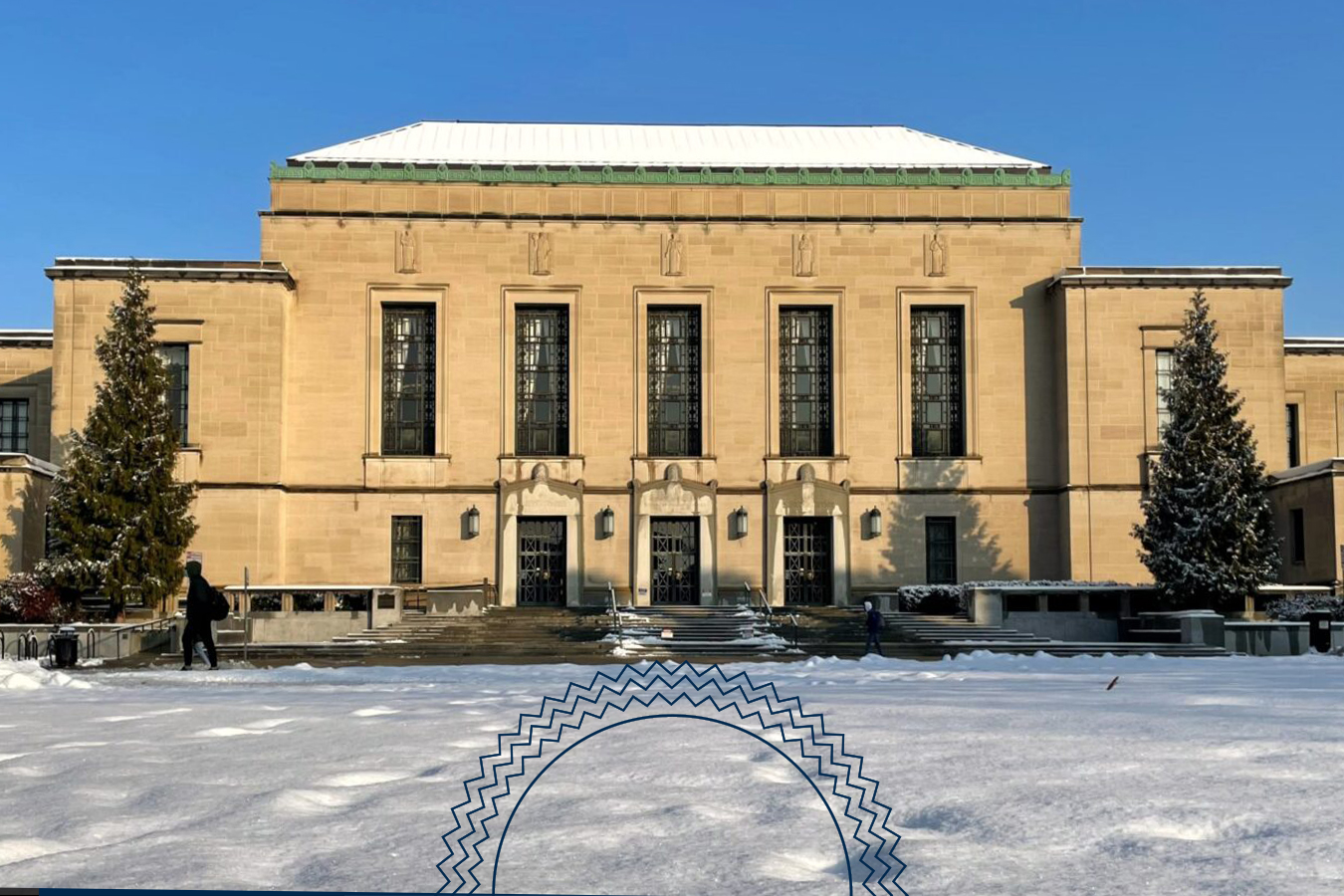 A large stone building with tall windows and two evergreen trees stands behind a snow-covered lawn under a clear blue sky.