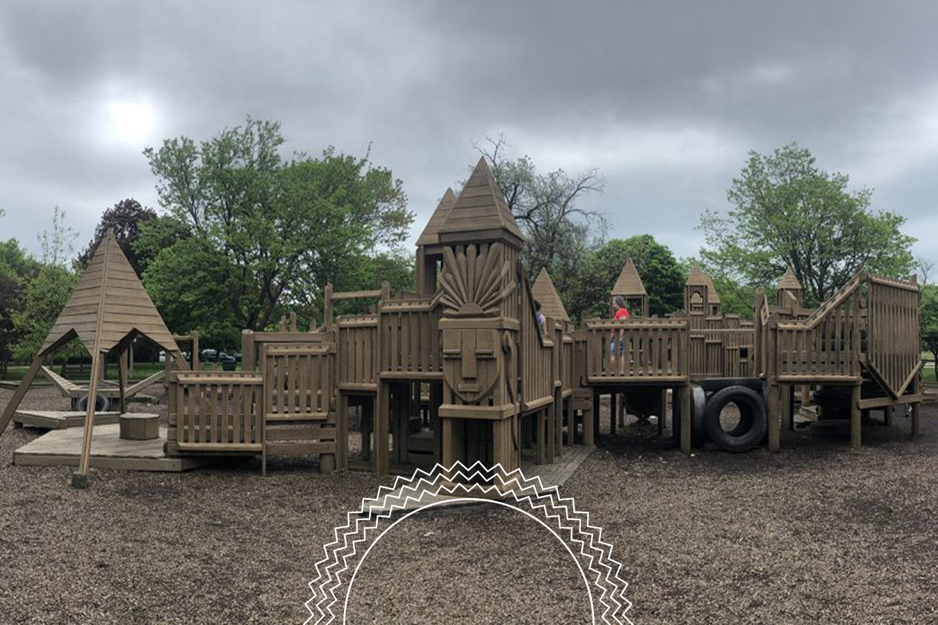 Large wooden playground structure with towers, ramps, and bridges, surrounded by trees under a cloudy sky; tire obstacle visible on the right.