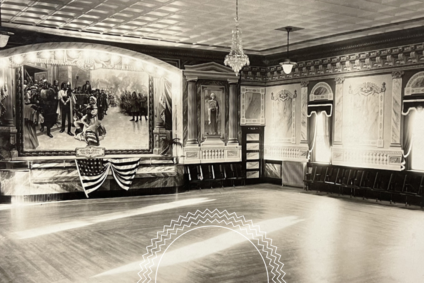 A vintage ballroom with ornate walls, a chandelier, rows of empty chairs, and a large mural above a stage decorated with an American flag bunting.
