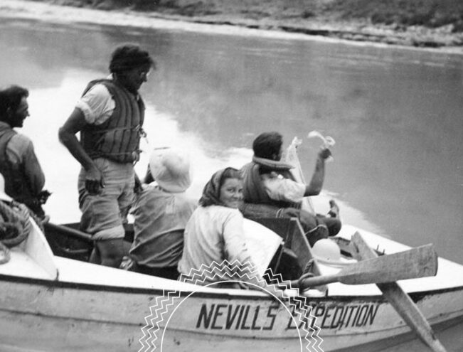 Six people in a small boat labeled “Nevills Expedition” are wearing life jackets and hats, with one person looking back and smiling, on a calm river near a grassy bank.