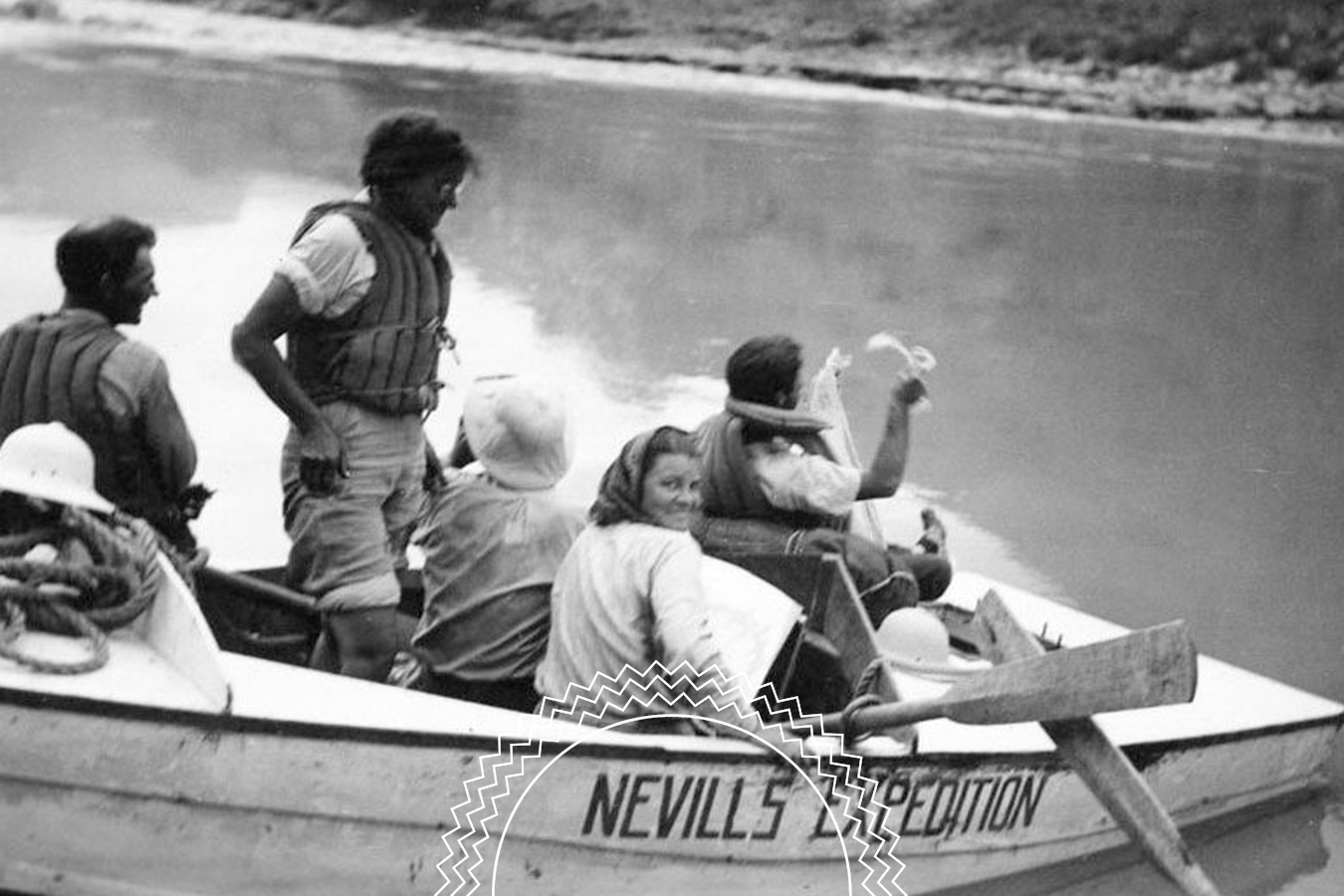 Six people in a small boat labeled “Nevills Expedition” are wearing life jackets and hats, with one person looking back and smiling, on a calm river near a grassy bank.