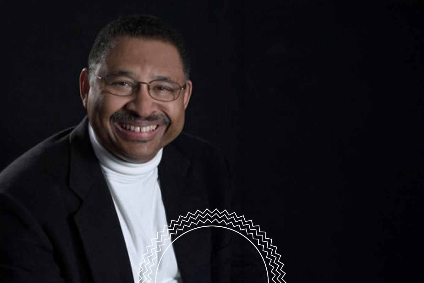 A man with glasses and a mustache smiles while wearing a white turtleneck and dark blazer, posed against a plain black background.