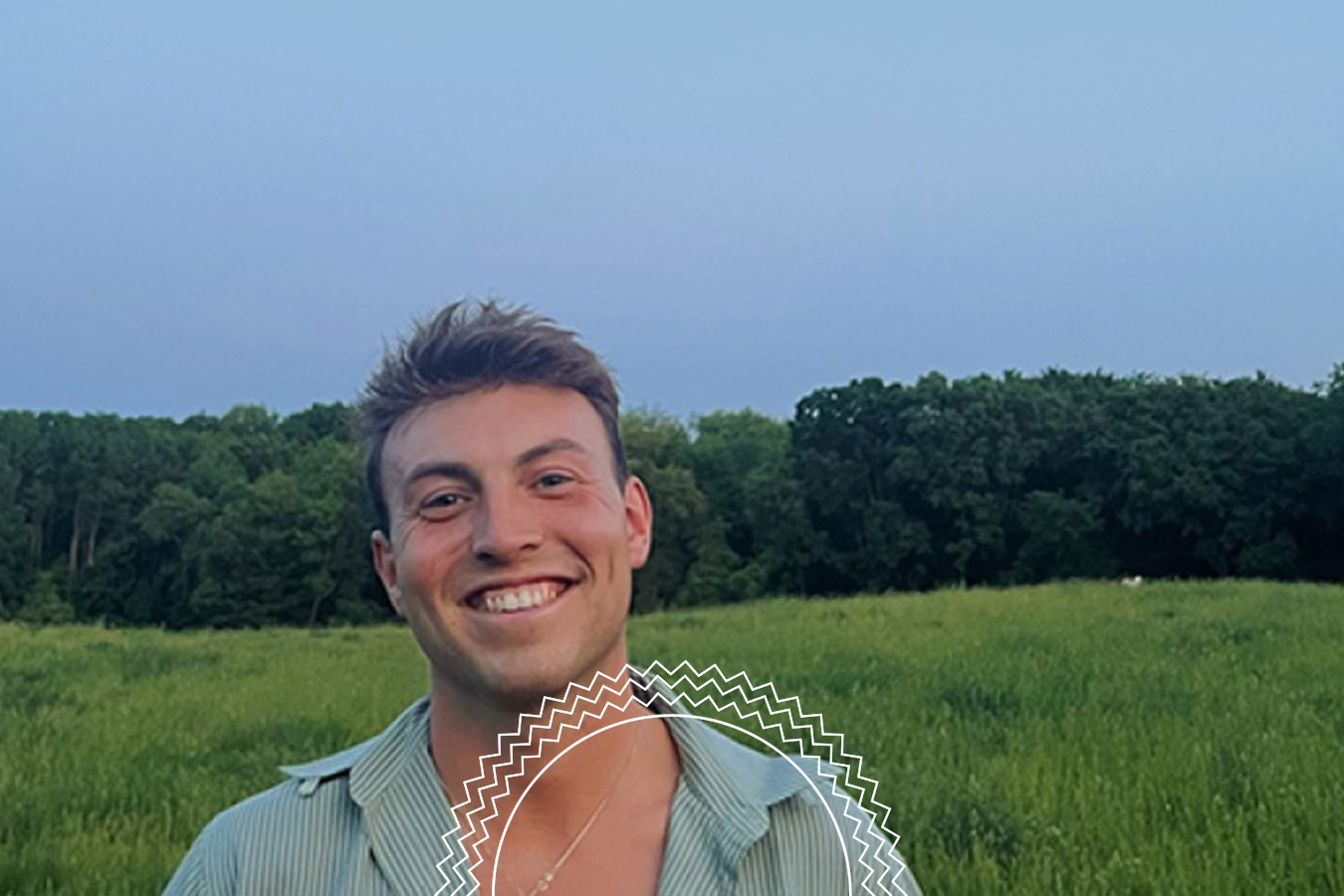A person stands smiling in a grassy field with trees in the background under a clear sky.