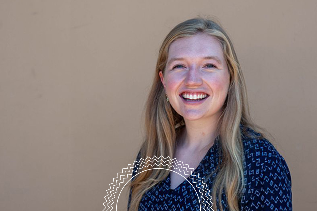 A person with long blonde hair smiles at the camera, wearing a dark patterned shirt, standing against a plain beige background.