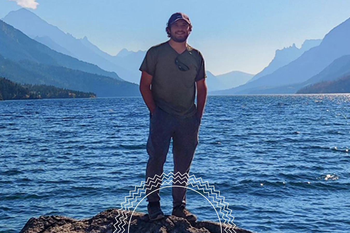A person stands on a rock by a large lake, surrounded by mountains under a clear blue sky.