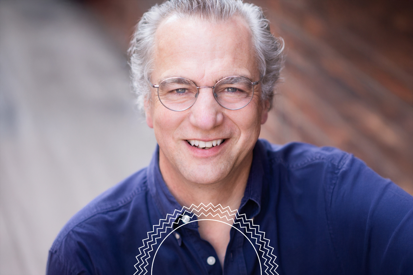 A middle-aged man with gray hair and glasses smiles at the camera, wearing a dark blue shirt.