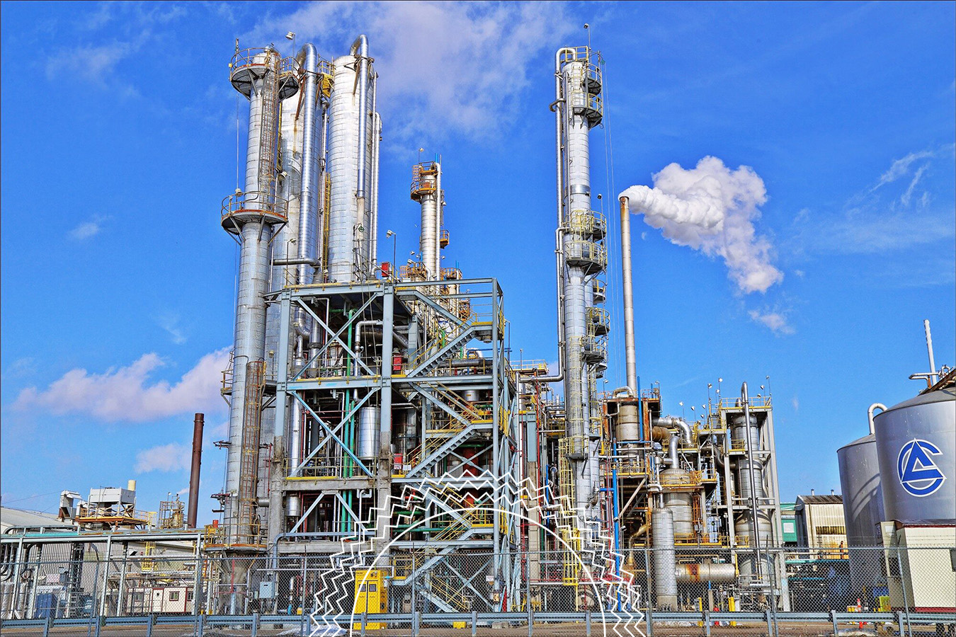 Industrial refinery plant with tall metal towers, pipes, and tanks, emitting steam into the blue sky, surrounded by a fenced area.