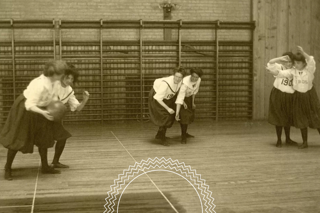 Four women in early 1900s athletic attire play basketball in a gymnasium with wooden floors and wall bars in the background.