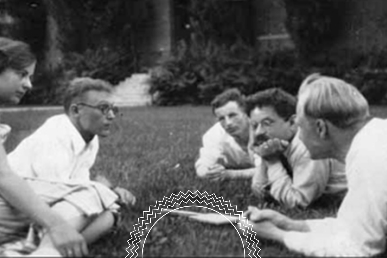 Four people sit and lie on grass in a circle, engaged in conversation, with trees and a building visible in the background.
