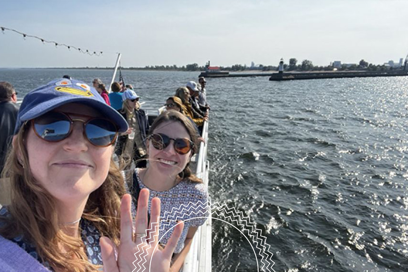 Two women take a selfie and wave while standing with others on a boat or ferry, with water and a distant shoreline visible in the background.