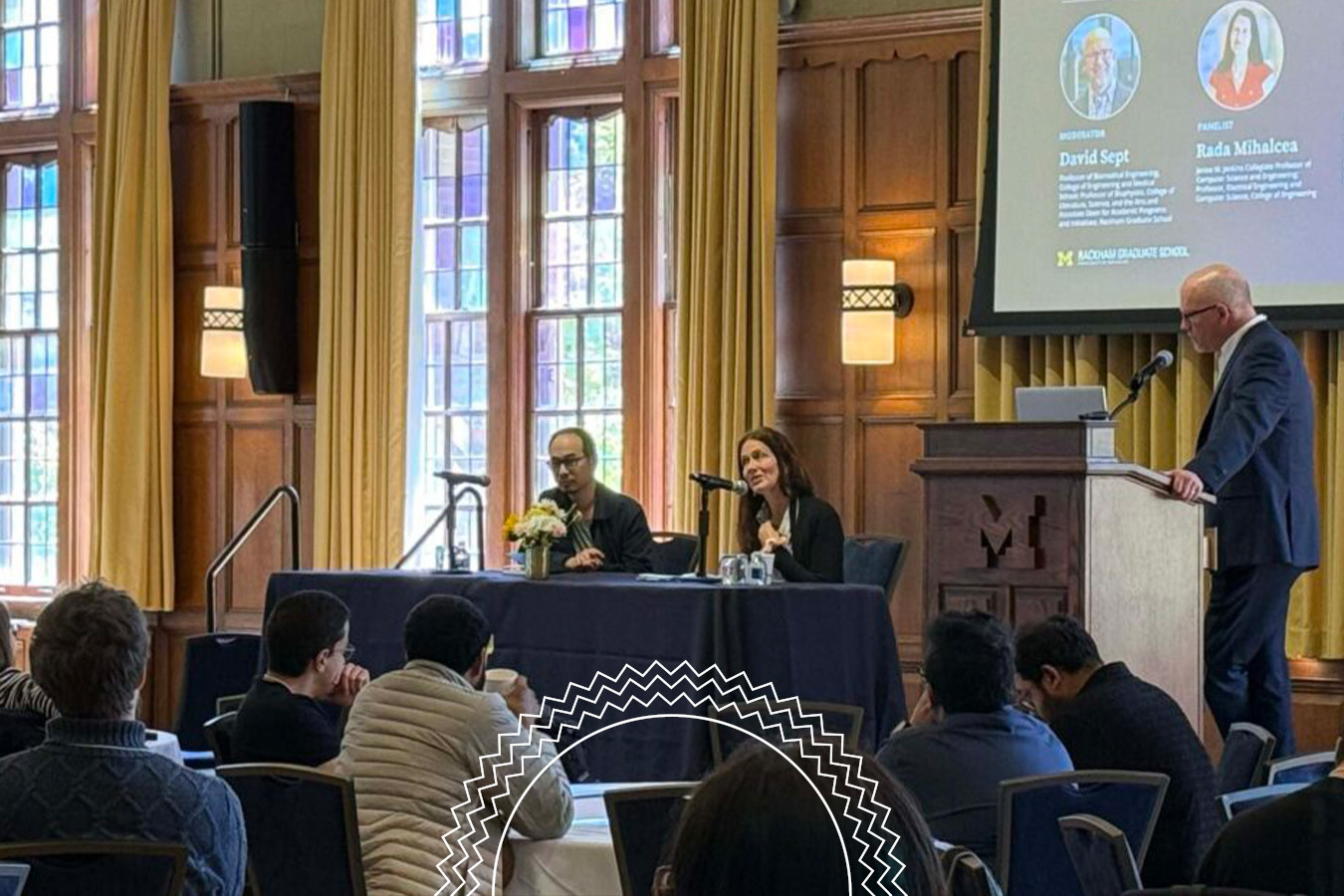 Two speakers sit at a table addressing an audience in a wood-paneled room, while a moderator stands at a podium near a screen with speaker information.