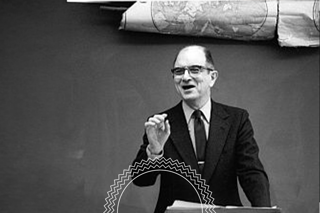 A man in a suit speaks at a lectern in front of a blackboard and a rolled-up map.