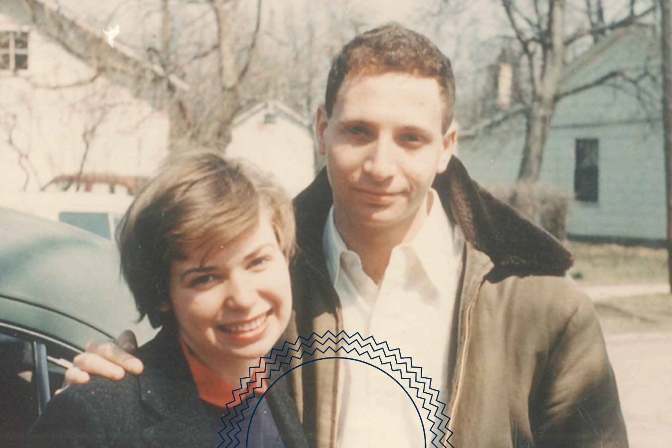 A young man and woman stand outside together, smiling at the camera. The background shows houses, leafless trees, and a parked car.