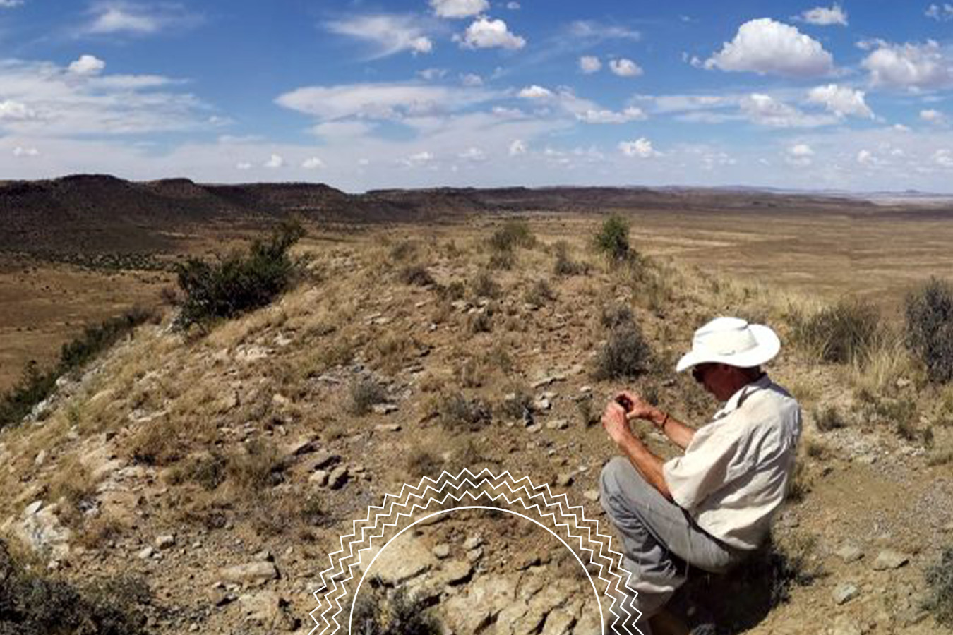 A person in a white hat examines rocks on a dry, rocky hillside under a partly cloudy sky, with expansive plains and distant mesas in the background.