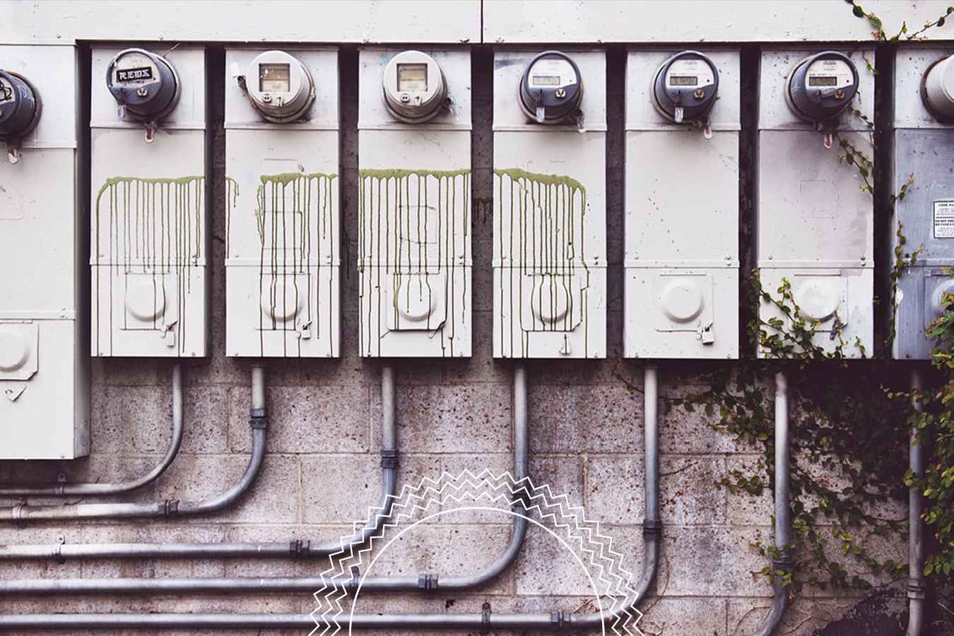 A row of electrical meters mounted on a wall, some with green paint drips. Metal pipes run beneath. Ivy grows along the right side.