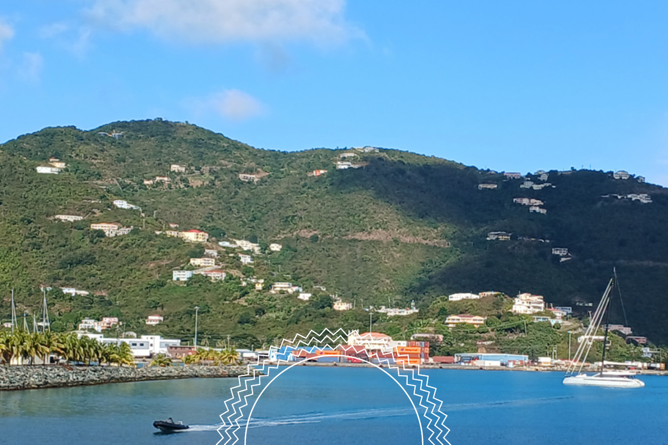 A small motorboat creates a wake on blue water near a coastal town, with green hills and scattered houses in the background under a blue sky.