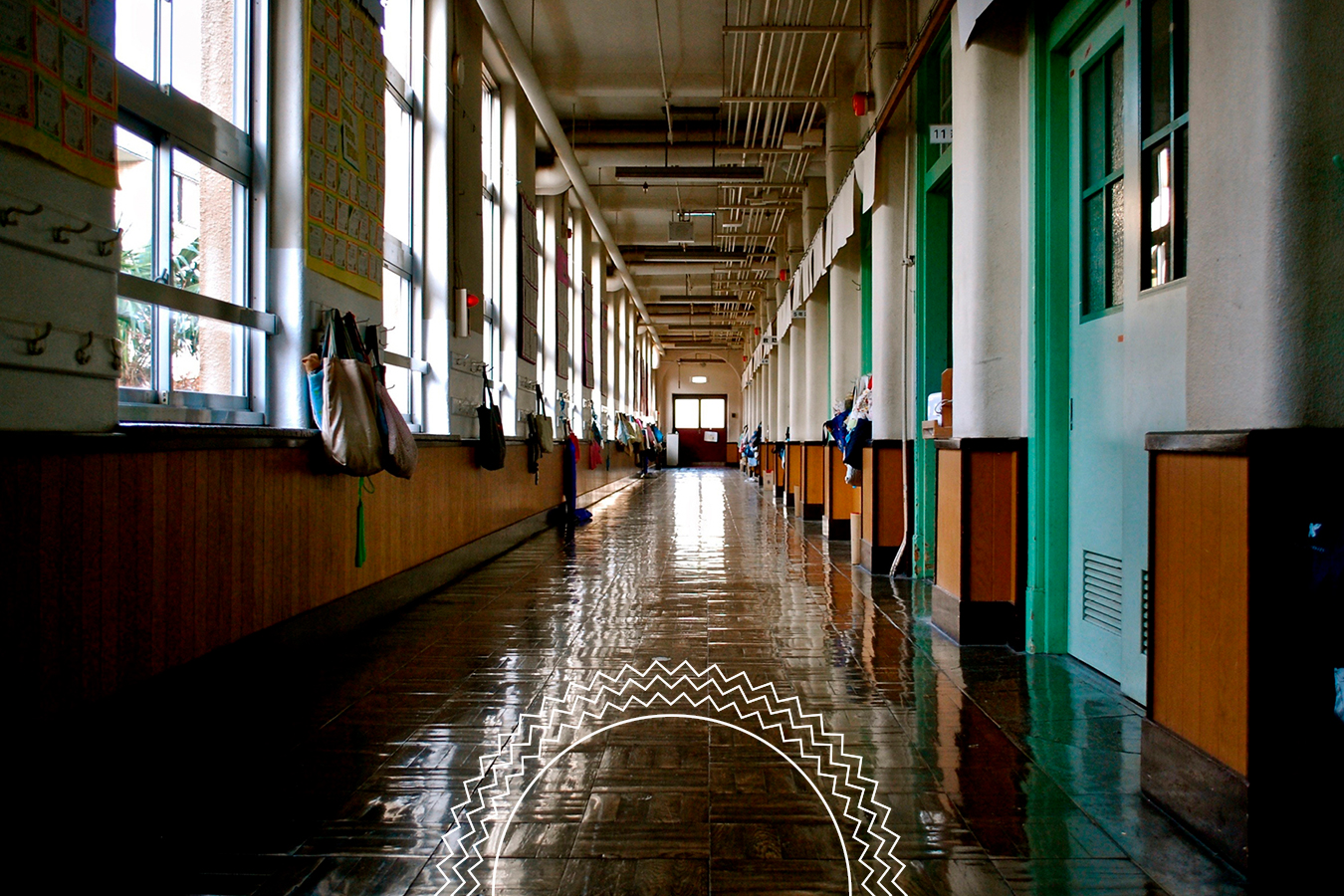 Long, empty school hallway with polished floors, green doors, windows on one side, and bags hanging along the wall.