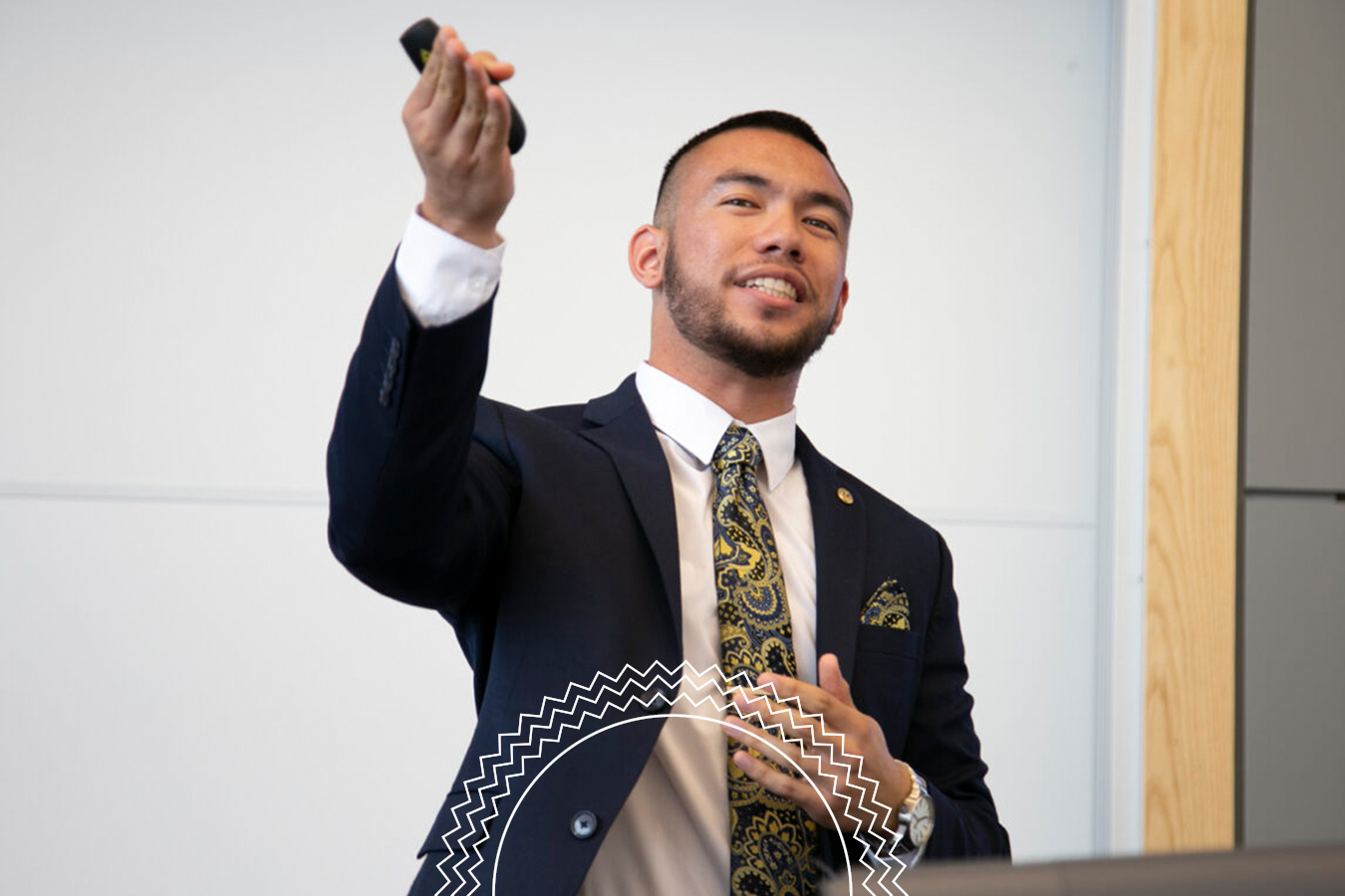 A man in a suit stands and gestures during a presentation, holding a remote control in one hand.