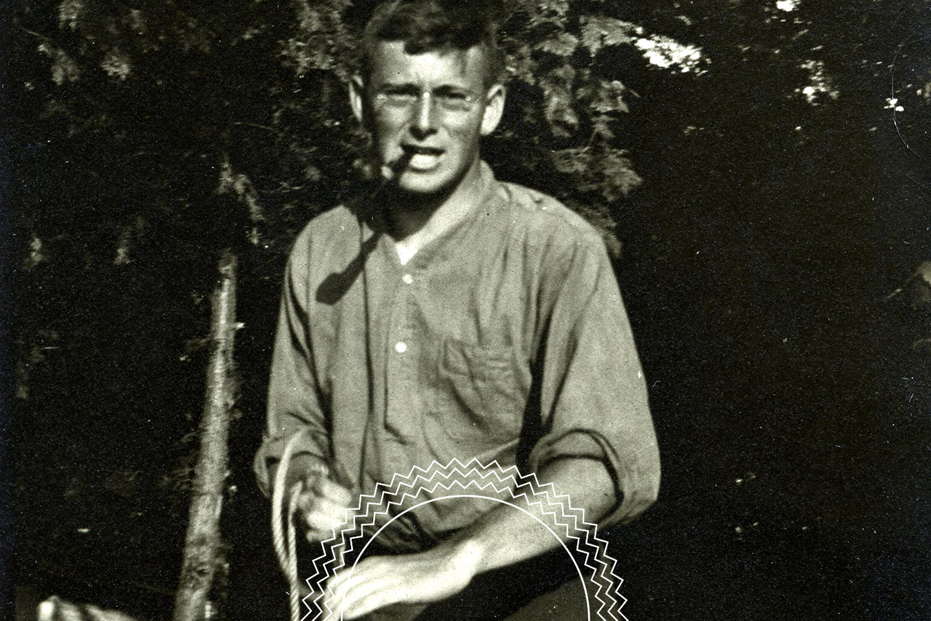 A young man with glasses and a pipe sits outdoors in front of trees, wearing a long-sleeved shirt, with hands resting on his lap.