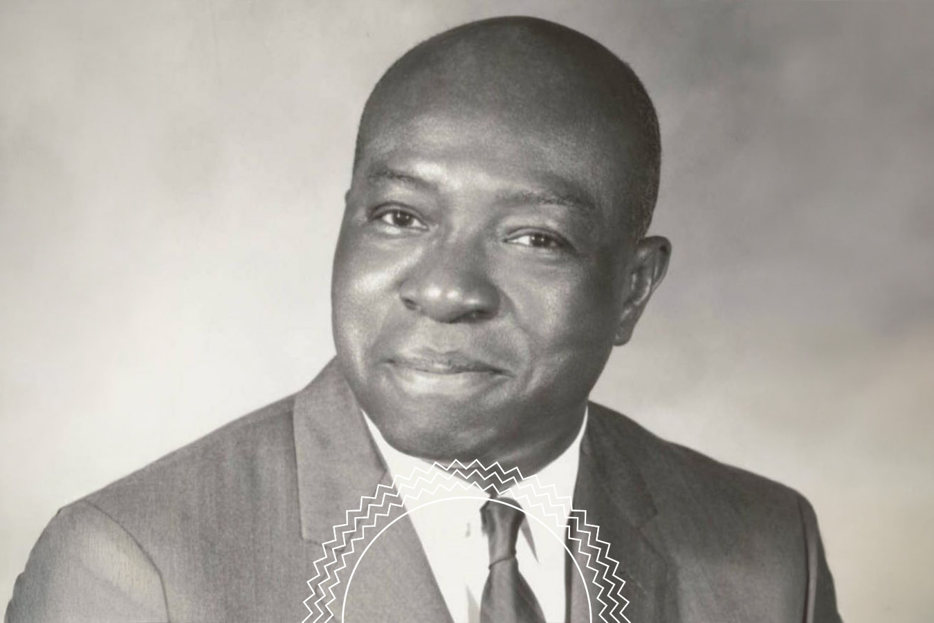 Black-and-white portrait of a man in a suit, looking at the camera and smiling slightly, with a neutral background.