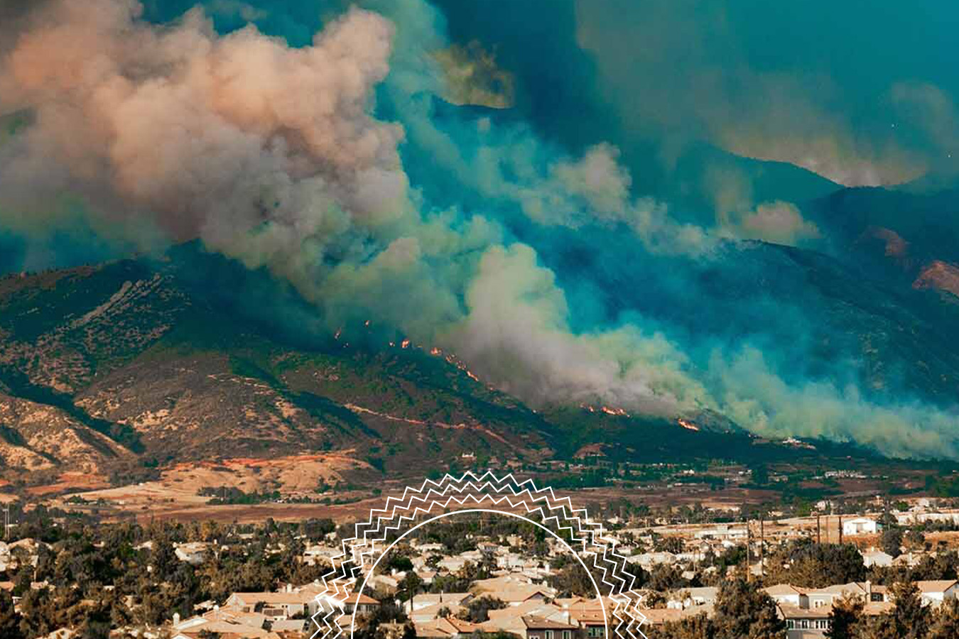 Wildfire burns on a mountain slope, producing thick smoke that drifts over a residential neighborhood in the foreground.