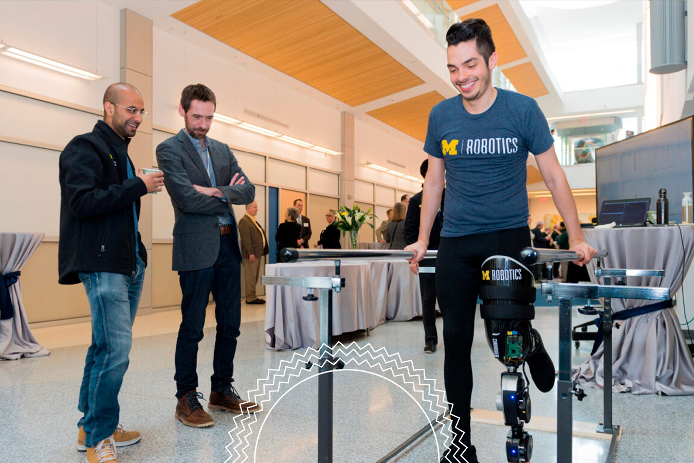 A man with a prosthetic leg, branded “M Robotics,” demonstrates walking with parallel bars as two men observe in a modern indoor setting.
