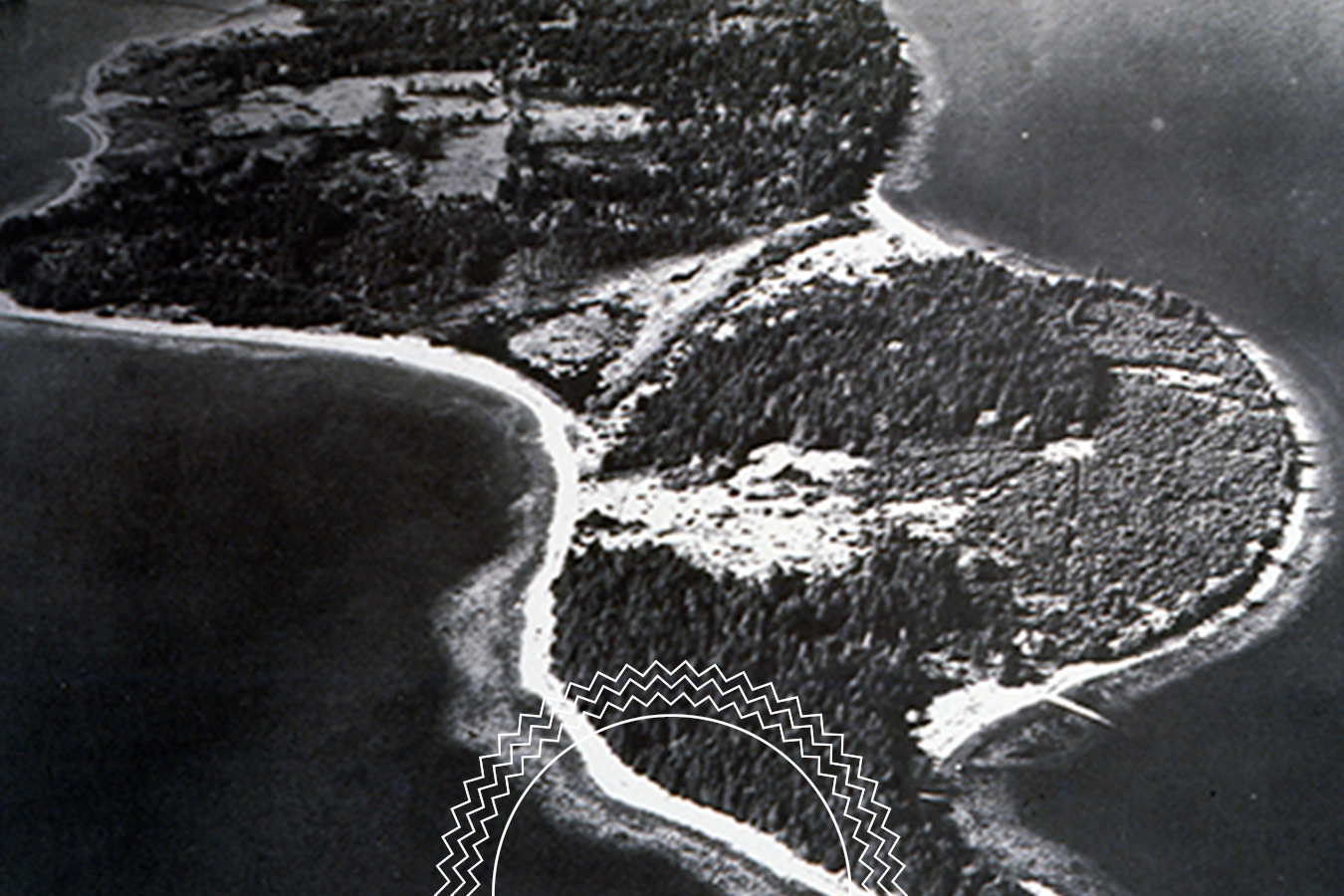 Aerial black and white photo of an island with visible tree cover, shoreline, and a narrow land bridge connecting two larger land masses, surrounded by water.