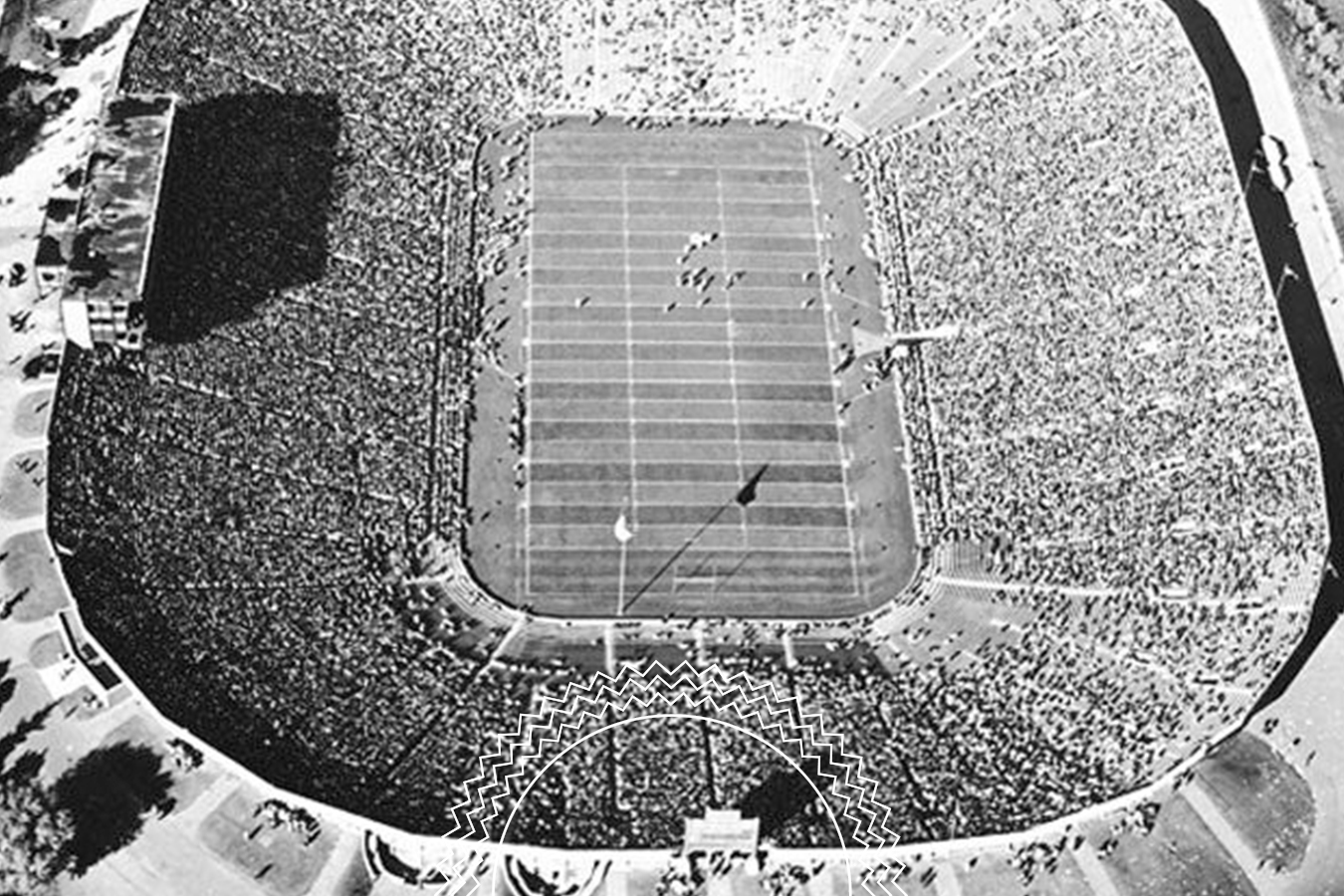 Black-and-white aerial view of a packed football stadium during a game, with spectators filling the stands and players visible on the field.