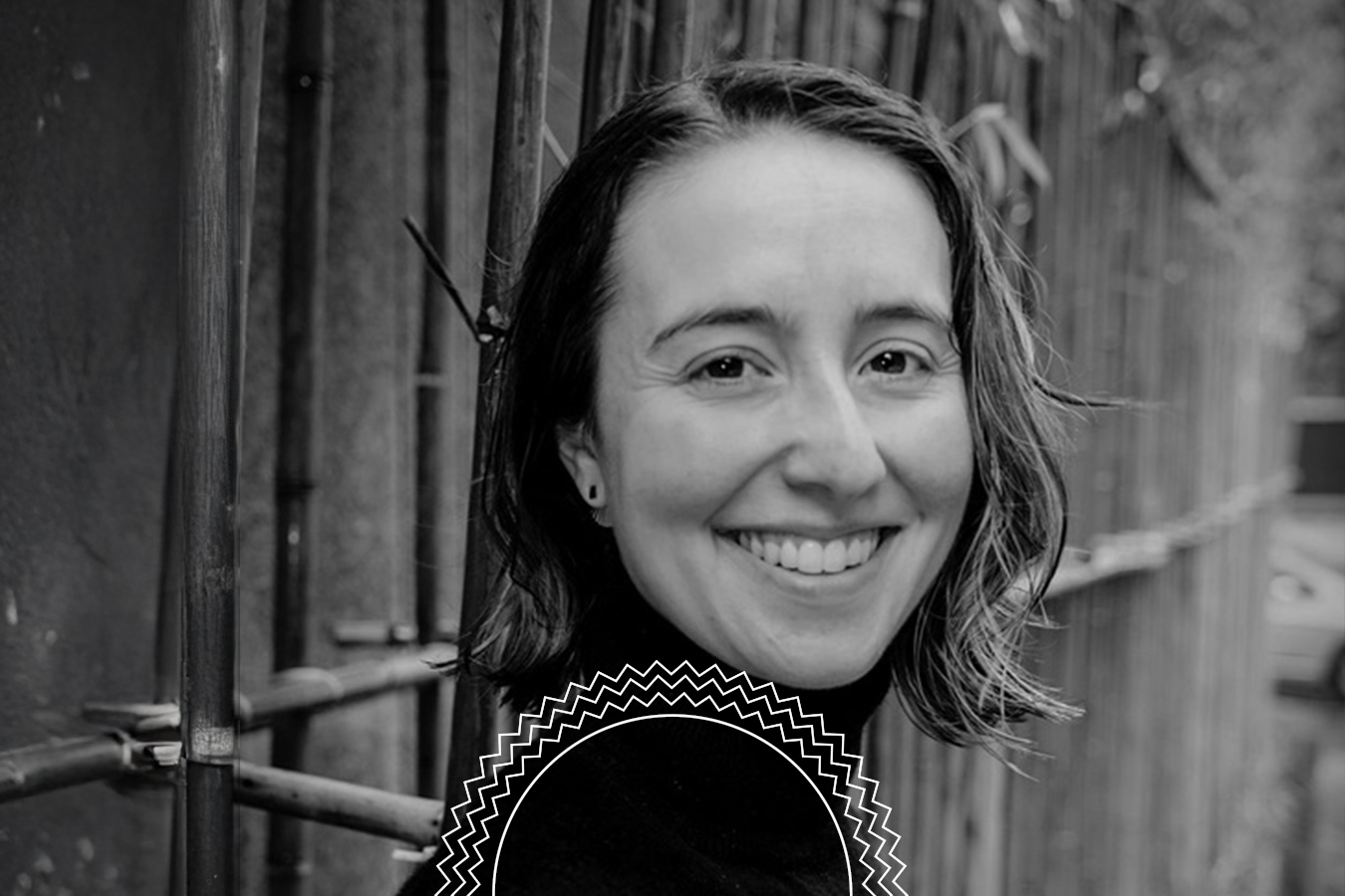 A woman with shoulder-length hair smiles at the camera while standing in front of a metal fence outdoors.