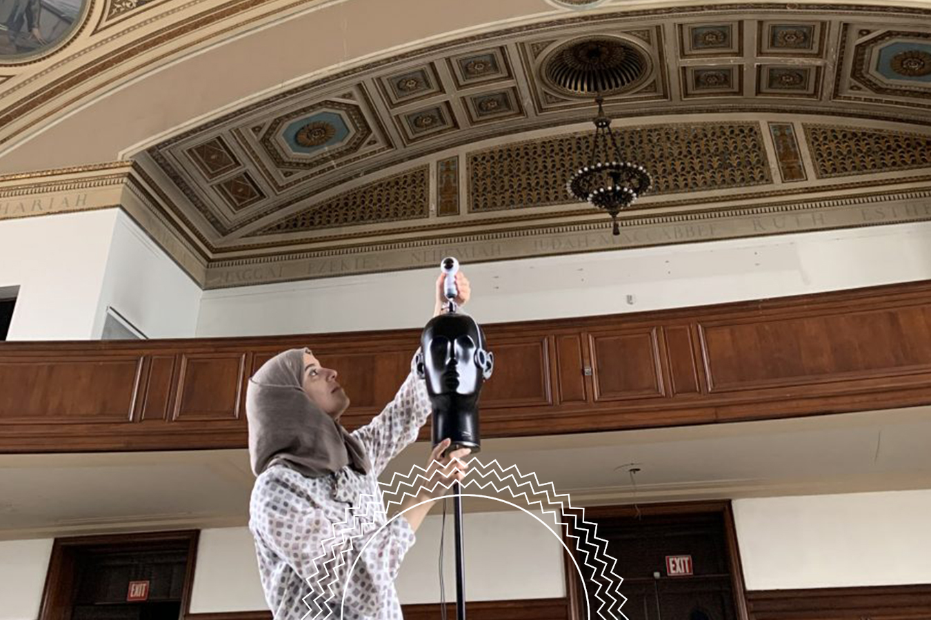 A person in a patterned shirt and headscarf adjusts a mannequin head with attached microphone inside a large, ornate room with a decorated ceiling.