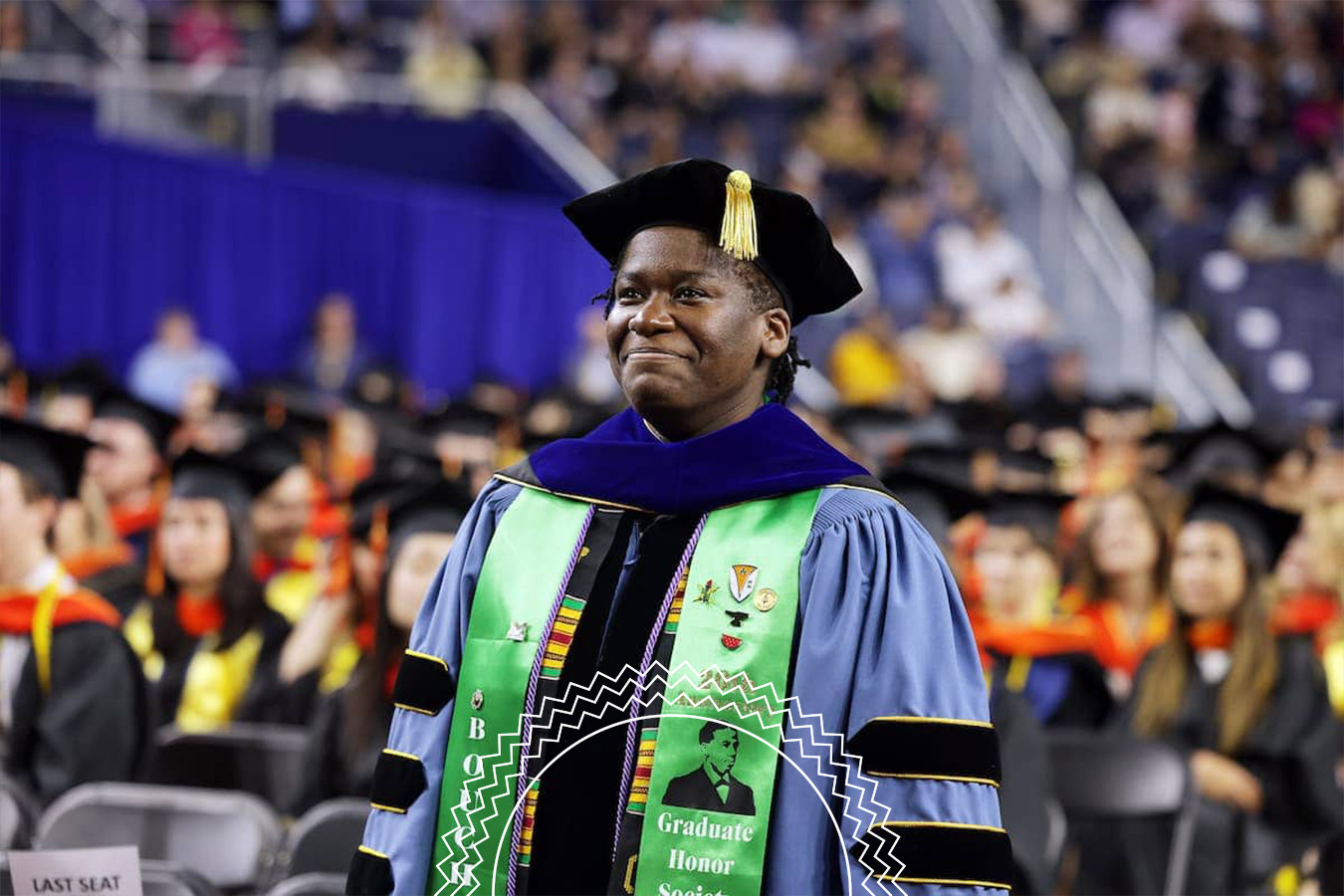 A graduate in academic regalia stands and smiles at a commencement ceremony, with an audience and other graduates seated in the background.