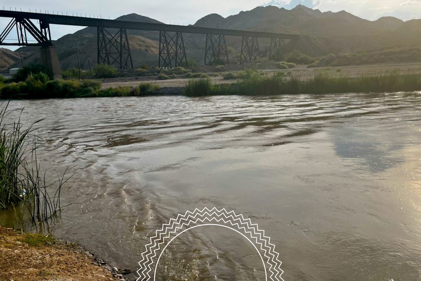 A wide river with gentle ripples flows in the foreground; a tall, metal railway bridge spans across hills in the background under a partly cloudy sky.