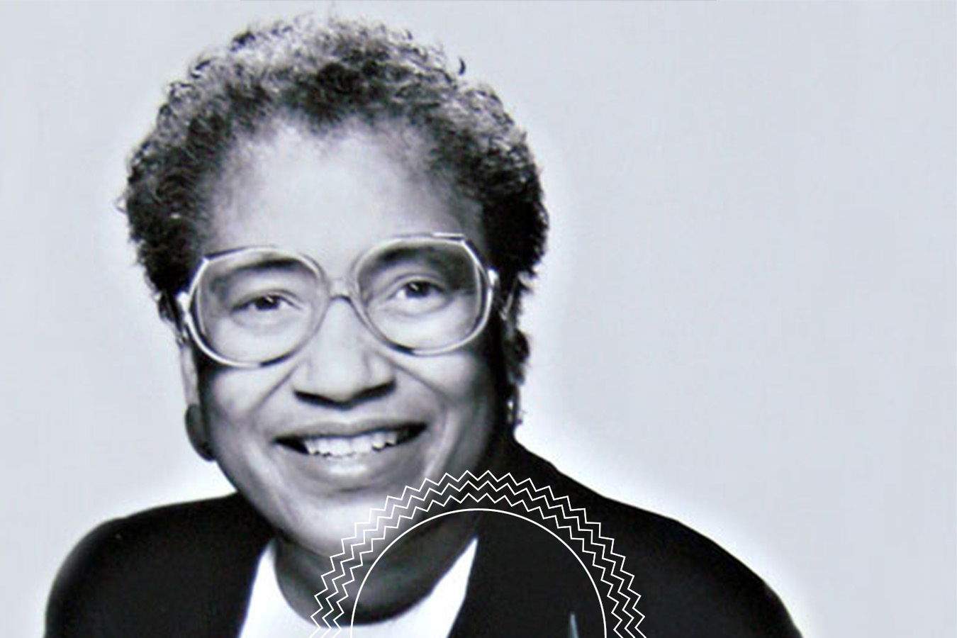 Black and white portrait of an older person with short curly hair, glasses, and a suit jacket, smiling at the camera. There is a partial circular decorative element at the bottom.