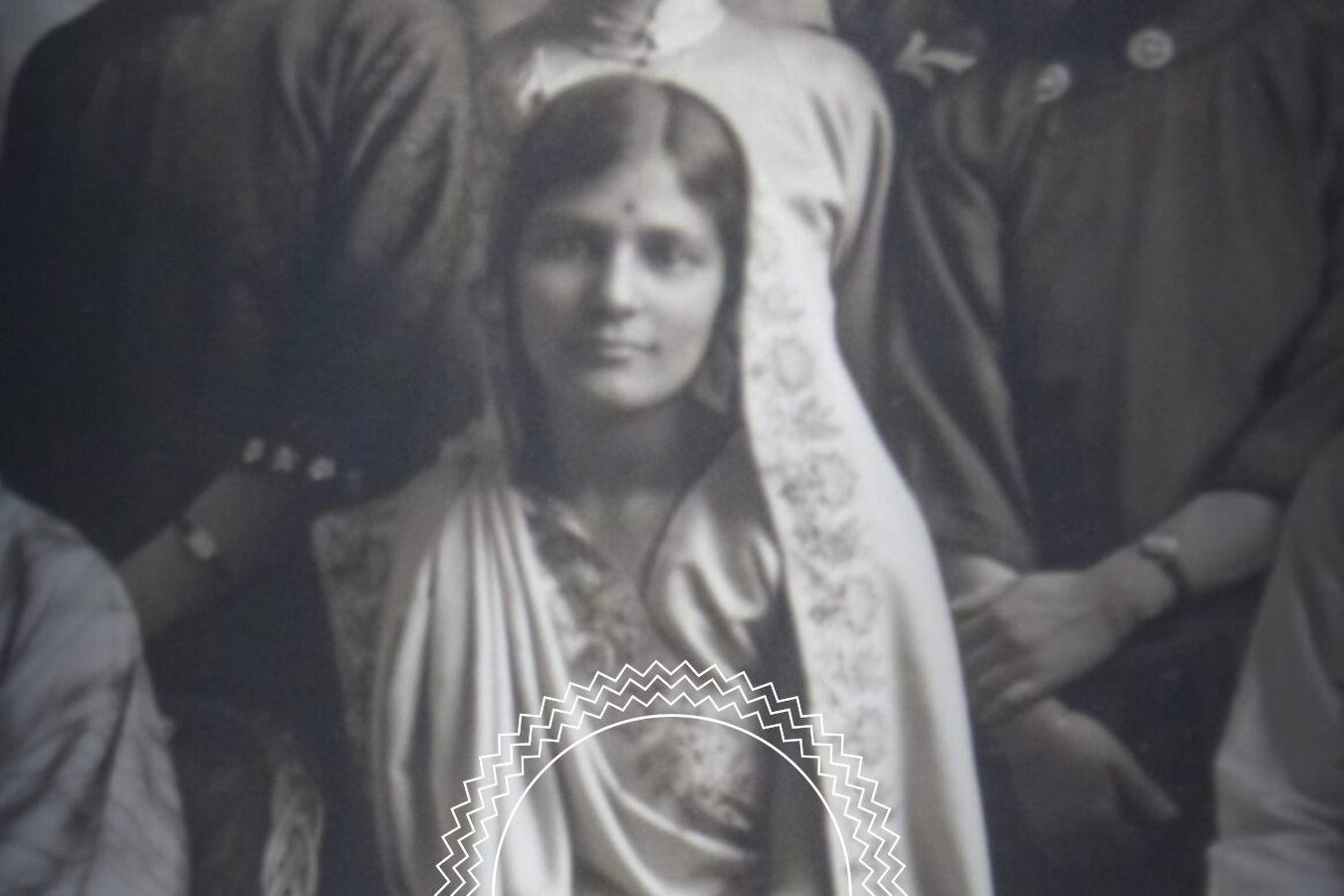 A woman in traditional attire with a bindi on her forehead sits in front of a group of people, looking at the camera.