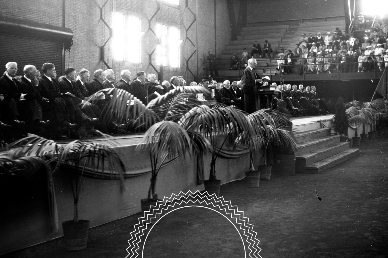 A speaker addresses an audience from a stage lined with potted palms, while people in formal attire are seated on the stage and in bleachers.