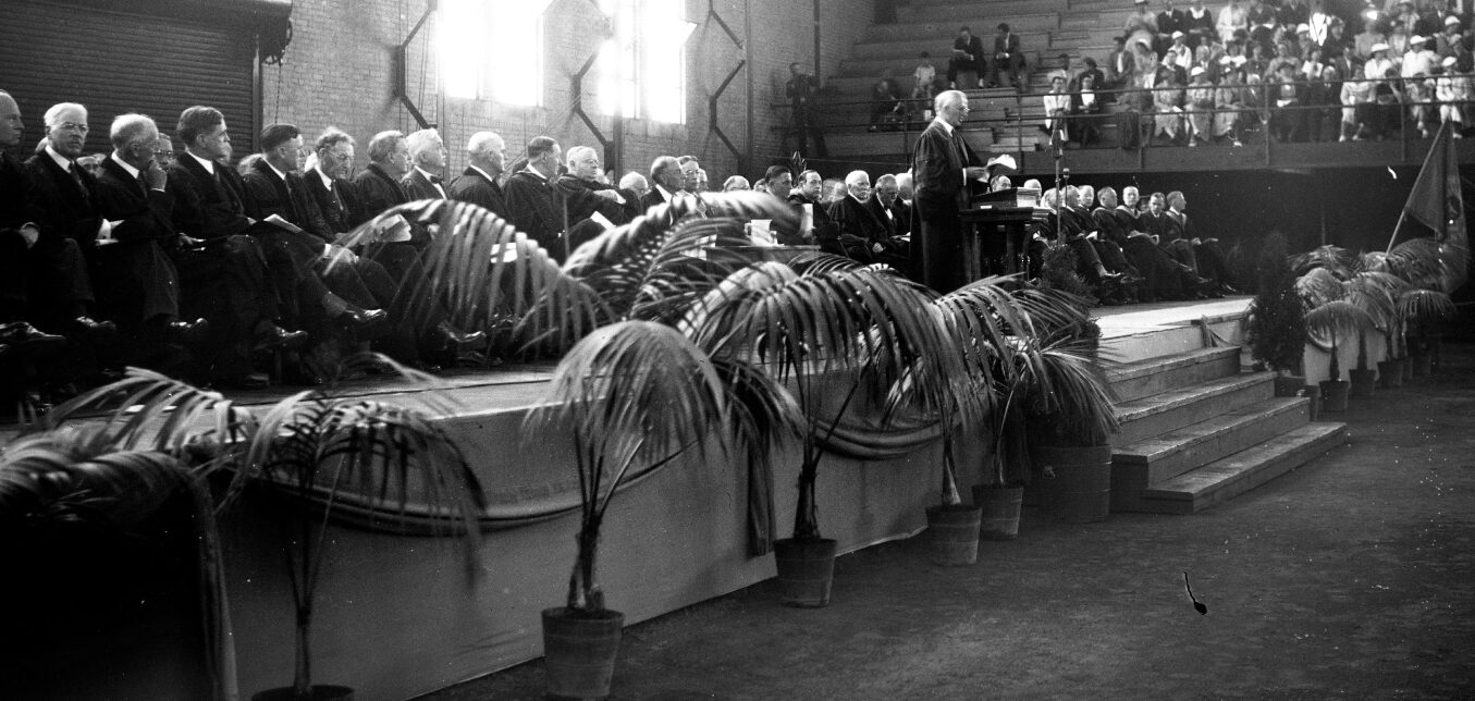 A speaker addresses an audience from a podium on a stage lined with potted plants, with seated officials and spectators in the background of a large indoor venue.