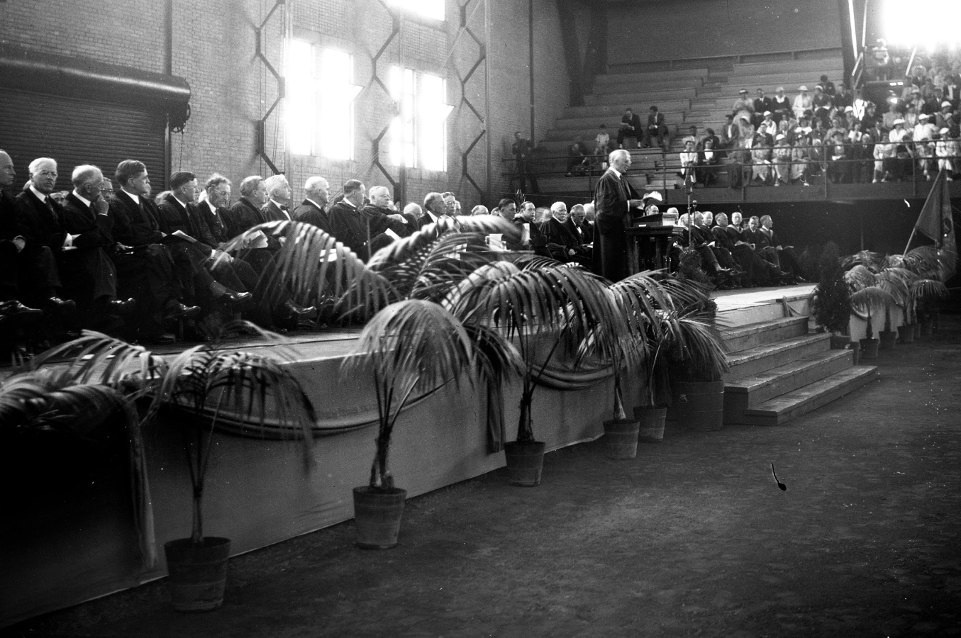 A speaker addresses an audience from a podium on a stage lined with potted plants, with seated officials and spectators in the background of a large indoor venue.