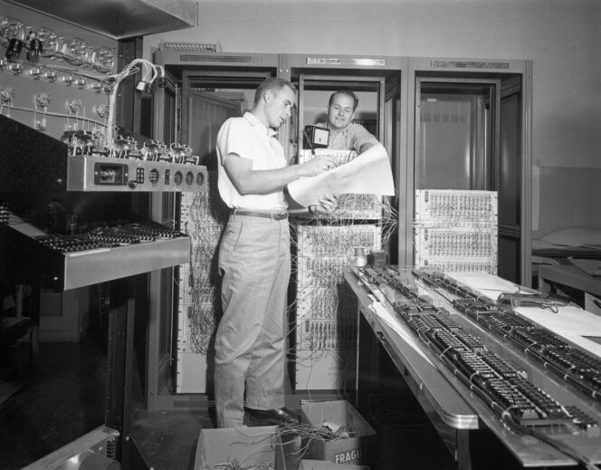 Two men examine large diagrams and control panels in a room filled with vintage computer equipment, cables, and circuit boards.