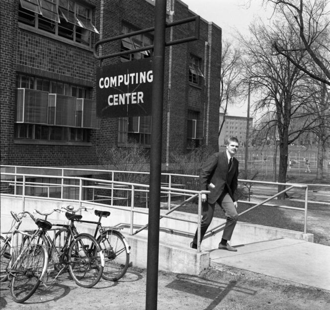 A man in a suit walks down steps outside a building labeled “Computing Center.” Three bicycles are parked nearby, and leafless trees are visible in the background.